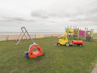 A playground with swings slide tunnel and climbing structures on grass by the beach at Shore Thing 280 in Wilsthorpe