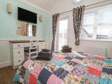A bedroom with a bed covered in a patchwork quilt and folded towels on top a desk with a chair a wall-mounted television and windows with stone pattern curtains at Shore Thing 279 in Wilsthorpe