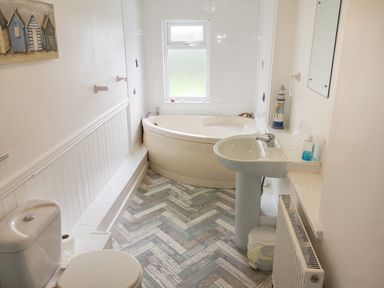A bathroom with a corner bathtub, toilet, pedestal sink, radiator, and wall art at Gull Cottage in Tintagel