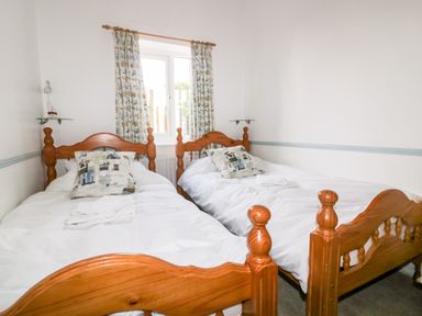 A bedroom with two wooden single beds white bedding patterned pillows and a window with curtains at Gull Cottage in Tintagel