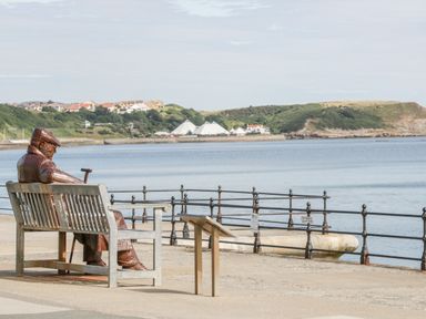 A statue of a person sitting on a bench facing a body of water with railings and hills in the background at Royal Victoria House in Scarborough