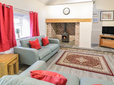 A living room with grey sofas and red cushions a brick fireplace with a wood stove a red patterned rug and a television at Crud yr Awel Braichmelyn near Bethesda