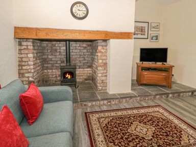 A living room with a brick fireplace and burning wood stove a sofa with red cushions a patterned rug a wooden TV stand with a TV and framed pictures on the wall at Crud yr Awel in Braichmelyn near Bethesda