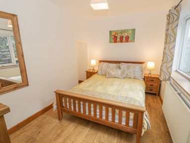 A bedroom with a wooden bed with yellow and grey bedding two bedside tables with lamps a window with patterned curtains a large wooden framed mirror and a painting of flowers at Crud yr Awel in Braichmelyn near Bethesda