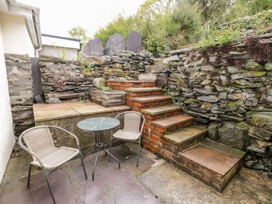 An outdoor patio with a round glass table and two woven chairs next to brick steps and stone walls at Crud yr Awel in Braichmelyn near Bethesda