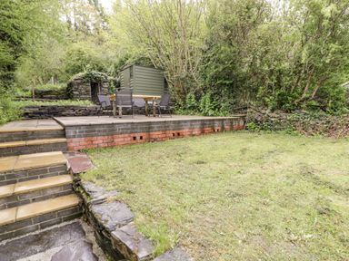 A garden with a grass lawn stone steps a raised patio table and chairs a shed and trees at Crud yr Awel in Braichmelyn near Bethesda