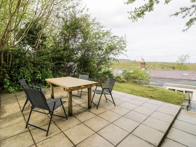 A patio with a wooden table and four black chairs surrounded by trees and overlooking rooftops and greenery at Crud yr Awel in Braichmelyn near Bethesda