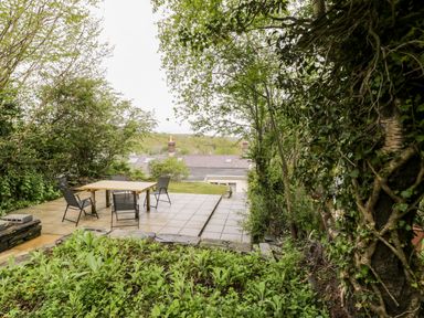 An outdoor patio area with a wooden table and four chairs surrounded by trees and plants at Crud yr Awel in Braichmelyn near Bethesda