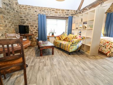 A living room with a wooden dining table and chairs a colorful sofa a wooden coffee table a shelving unit and blue curtains at The Forge at Smithy Cottage in Bloxham
