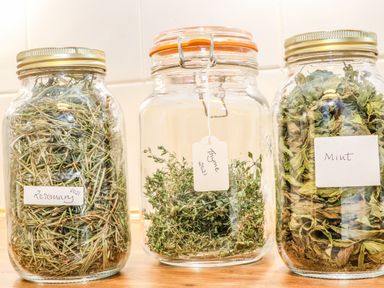 Three glass jars on a wooden surface containing dried rosemary thyme and mint labeled with handwritten tags