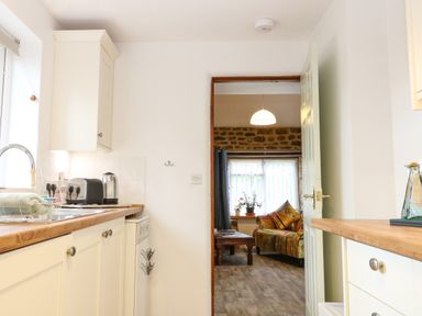 A kitchen with white cabinets, wooden countertops, a kettle, toaster, and a view into a living room with a patterned sofa and coffee table at The Forge at Smithy Cottage in Bloxham