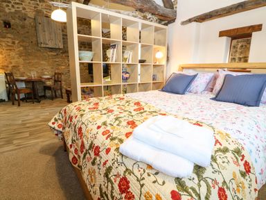 A bedroom with a floral bedspread and folded towels on the bed a wooden shelving unit and dining table with chairs against a stone wall at The Forge at Smithy Cottage in Bloxham