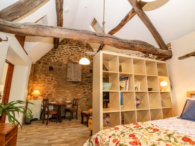 A bedroom with a wooden bed and a floral bedspread separated by shelving from a dining area with a wooden table and chairs in a room with exposed wooden beams and a stone wall at The Forge at Smithy Cottage in Bloxham