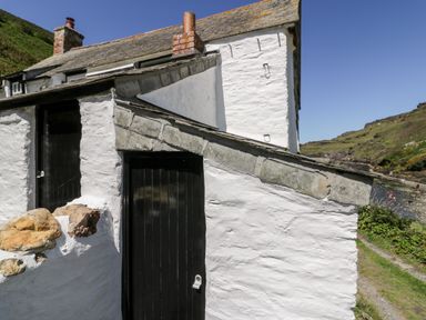 A white stone cottage with black doors and a sloped stone roof near a rocky hillside and water at Harbour Cottage in Boscastle