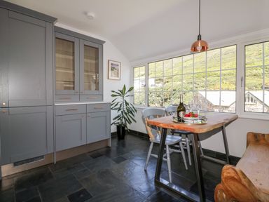 A dining area with a wooden table and chairs next to large windows and grey cabinets at Harbour Cottage in Boscastle