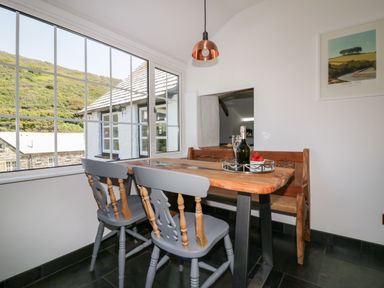 A dining area with wooden table wooden bench two chairs large window and a hanging light at Harbour Cottage in Boscastle