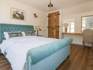 A bedroom with a teal upholstered bed a wooden floor a wooden double door a desk with a lamp and chair at Harbour Cottage in Boscastle