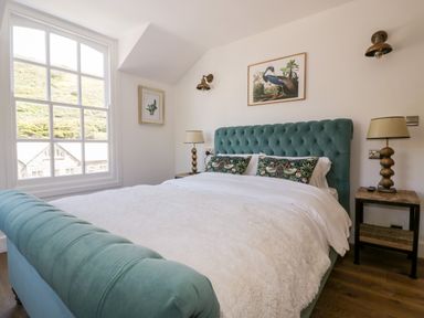 A bedroom with a large bed with teal tufted headboard and footboard two bedside tables with lamps and framed pictures on the walls at Harbour Cottage in Boscastle