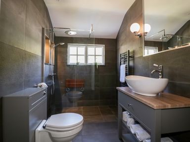 A bathroom with a toilet, sink on a wooden countertop, shower area with a glass divider, towel rack, and window at Harbour Cottage in Boscastle