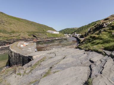 A coastal scene with a stone pier, grassy hills, and small buildings near a rocky shore at Harbour Cottage in Boscastle