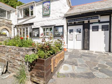 Exterior of a white building with signs for Museum of Witchcraft and Magic and a garden with flowers at Harbour Cottage in Boscastle