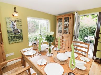 A dining room with a wooden table set for six people with plates glasses and green napkins beside a cabinet and an open door leading outside at Prospect Cottage in Malvern Wells near Great Malvern
