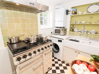 A kitchen with a stove and pots a washing machine a microwave on a counter and shelves with dishes and a sink at Prospect Cottage in Malvern Wells near Great Malvern