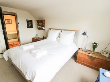 A bedroom with a white bedspread, two bedside tables, a wooden chest with a lamp and flowers, and a door leading to a bathroom at Prospect Cottage in Malvern Wells near Great Malvern