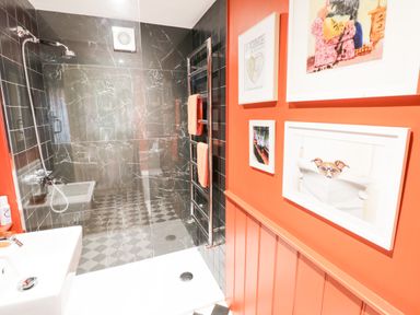 A bathroom with a walk-in shower with black tiles and checkered floor a white sink orange walls with framed pictures and a towel rack with orange towels at Prospect Cottage Malvern Wells near Great Malvern