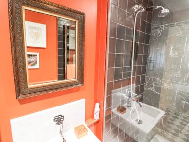 A bathroom with a shower area with black tiles and a glass door a sink with toiletries and a mirror on an orange wall at Prospect Cottage in Malvern Wells near Great Malvern