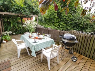 An outdoor wooden deck with a table covered by a polka dot tablecloth surrounded by white plastic chairs and a black charcoal grill nearby at Prospect Cottage in Malvern Wells near Great Malvern