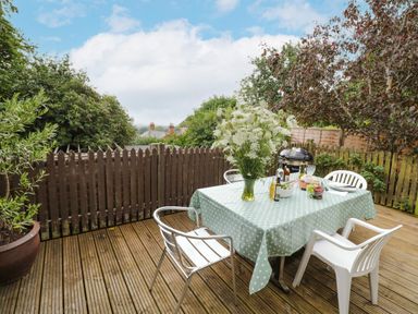 An outdoor wooden deck with a table covered by a polka dot tablecloth surrounded by chairs and a vase of flowers at Prospect Cottage in Malvern Wells near Great Malvern