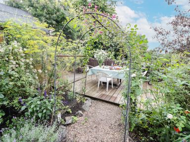 A garden pathway leading to a wooden deck with a table and chairs surrounded by plants and flowers at Prospect Cottage in Malvern Wells near Great Malvern