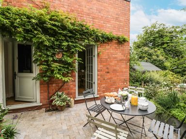 A patio with a black table and metal chairs and a table set for breakfast outside a brick house covered partially with green plants at Prospect Cottage in Malvern Wells near Great Malvern
