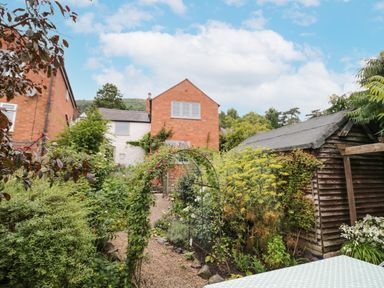 A garden with lush plants and flowers surrounding a gravel pathway and a wooden shed near a brick house in Malvern Wells near Great Malvern