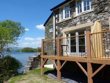 A stone house with a wooden balcony overlooking a lake and trees at Lowe McConnell in Far Sawrey