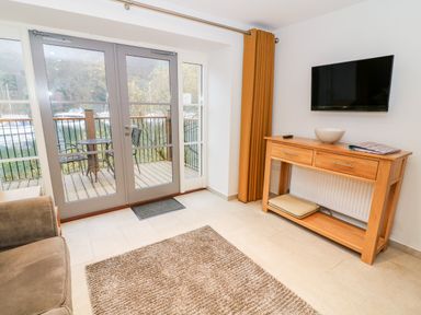 A living room with glass doors opening to a balcony with metal chairs and table a wooden console table below a mounted TV with a remote and bowl on it and a brown rug on the floor at Lowe-McConnell in Far Sawrey