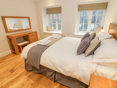 A bedroom with a double bed, wooden dressing table with mirror, two windows with cushions on the sills at Lowe-McConnell in Far Sawrey