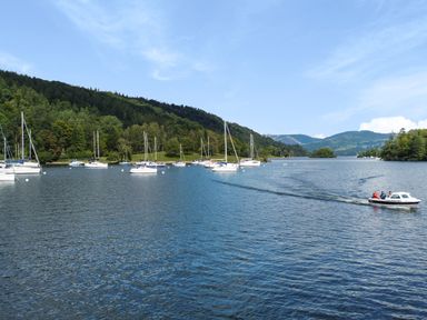 A lake with sailboats docked near the shore and a small motorboat moving on the water with hills and trees in the background at Lowe-McConnell in Far Sawrey