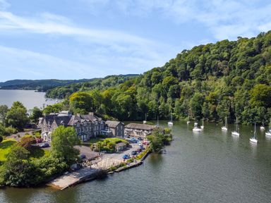 A waterfront estate with parked cars and moored boats next to a forested hillside at Lowe-McConnell in Far Sawrey