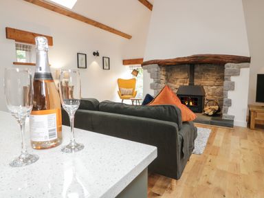A living room with a bottle and two glasses on a counter a dark sofa with orange cushions a yellow chair a stone fireplace and wood flooring at The Shippon in Llanrwst