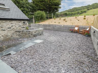 An outdoor gravel patio area with stone and wooden walls two wooden chairs and a small table with plants around at The Shippon in Llanrwst