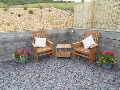 An outdoor seating area with two wooden chairs with white cushions a small wooden table and flower pots with red and pink flowers surrounded by wooden fencing