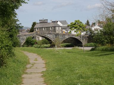 A stone arch bridge over a river with a paved path and green grass along the riverbank at The Shippon in Llanrwst