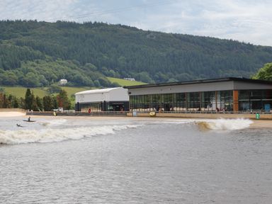 An artificial wave pool with surfers and buildings in front of a forested hillside at The Shippon in Llanrwst