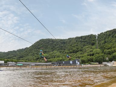 Two people zip lining over a body of water with green forested hills in the background at The Shippon in Llanrwst