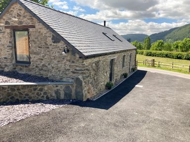 A stone building with a slate roof next to a paved driveway and green fields at The Shippon in Llanrwst