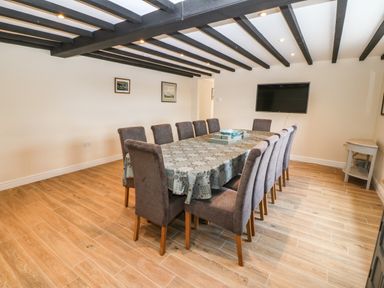 A dining room with a long table covered with a tablecloth and twelve chairs a wall-mounted television and wooden flooring at Trewen Botwnnog near Sarn Meyllteyrn