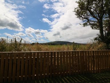 A wooden fence with tall grass behind it and a hill in the background under a partly cloudy sky at Trewen in Botwnnog near Sarn Meyllteyrn
