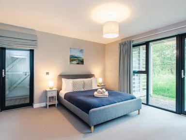 A bedroom with a bed with blue bedding two bedside tables with lamps and large glass doors at Wildcat Lodge in Feshiebridge near Aviemore